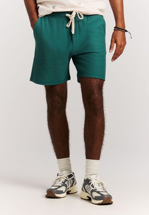 Mann trägt grüne Shorts mit Kordelzug, weiße Socken und graue und weiße Turnschuhe, steht mit einer Hand entspannt an der Seite.