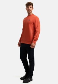 Sudadera naranja con cuello redondo y puños acanalados, combinada con pantalones negros y zapatillas negras. Presenta un pequeño logo en el pecho.