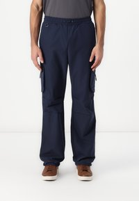 Homme portant un pantalon cargo bleu marine avec une ceinture élastique et des baskets marron, debout sur un fond blanc.