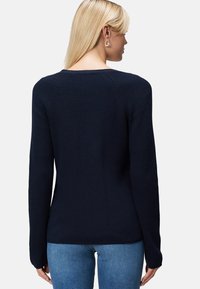 Femme aux longs cheveux blonds portant un pull en tricot bleu marine et un jean bleu clair, vue de dos sur un fond blanc.
