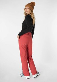 Pantaloni da sci rossi con vita alta e cerniere laterali, abbinati a un top nero a maniche lunghe e a un berretto lavorato a maglia marrone, indossati con stivali da sci bianchi e grigi.