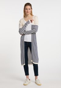 Cardigan lungo con apertura frontale in crema con righe navy e maniche aderenti. Abbinato a una maglietta bianca e jeans scuri, indossato con sneakers bianche.