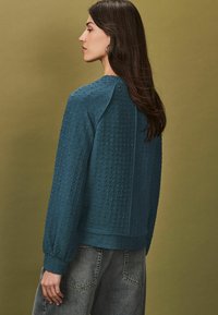 Teal gemêleerde sweatshirt met een ronde hals, geplooide mouwen en een relaxte pasvorm. Gecombineerd met lichtgrijze jeans, tegen een neutrale achtergrond.