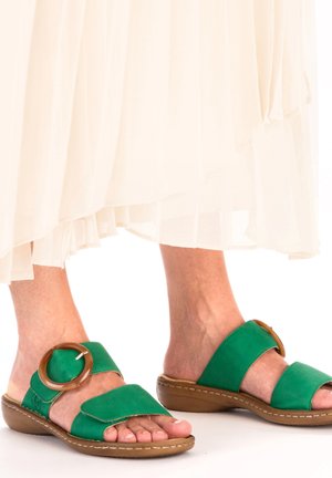 Sandales en cuir suédé vert avec de larges brides, un accent rond brun et une semelle basse beige, partiellement obscurcies par une jupe plissée blanche.