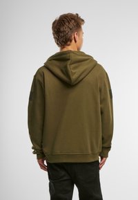 Felpa con cappuccio verde oliva, dal texture morbida, grande cappuccio e polsini a coste. Presenta una toppa sulla manica sinistra e una vestibilità rilassata.
