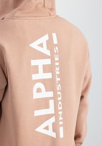 Hoodie beige chiaro realizzato in un tessuto morbido, con un grande logo bianco "ALPHA INDUSTRIES" allineato verticalmente sul retro.