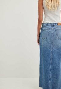 Jeans-maxiklänning i ljusblå med fransar i kanten, med två bakfickor och en märkesetikett på midjan. Ribbat vitt linne synligt.