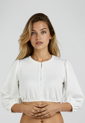 Junge Frau mit offenen blonden Haaren trägt eine weiße, strukturierte, gekürzte Bluse mit Puffärmeln vor einem schlichten grauen Hintergrund.