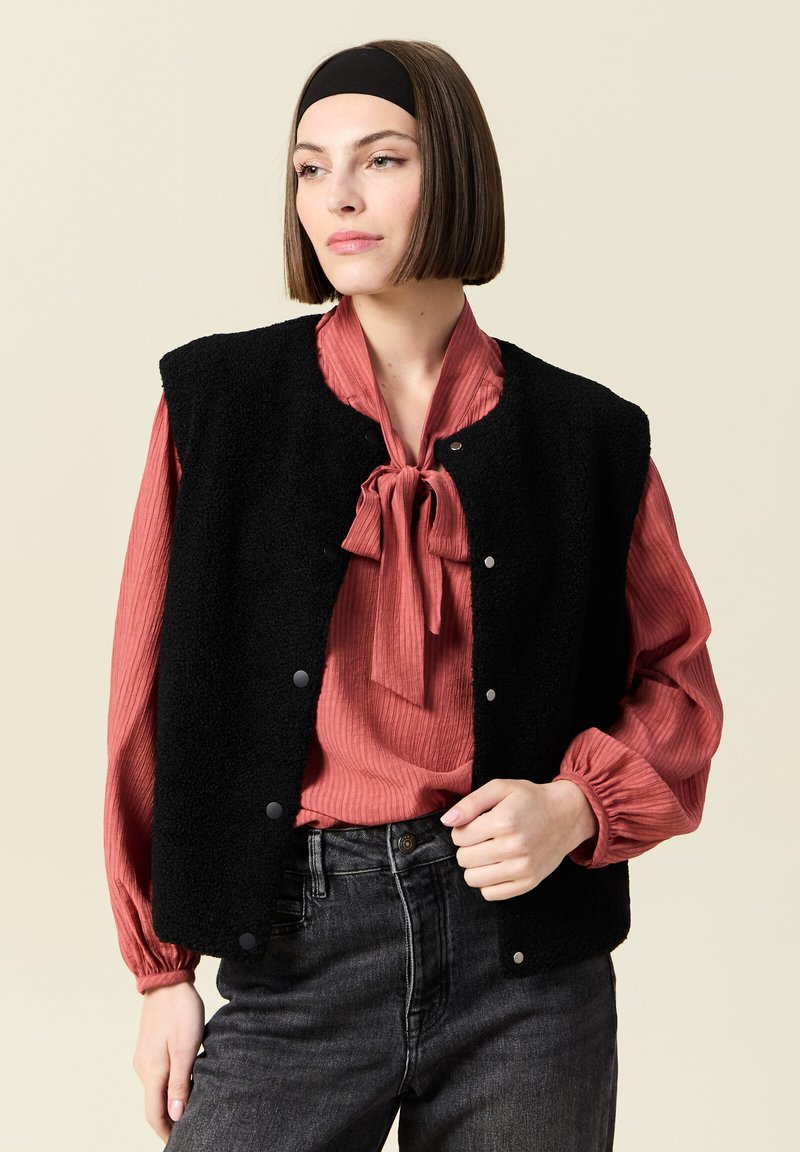 Svart fleeceväst med tryckknappar, bärs över en ribbad, röd blus med knytkrage. Modellen är stylad med mörka jeans och ett svart hårband.