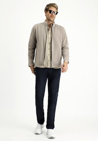 Hellbeige Fleece-Zip-Jacke, beiges Hemd mit Knöpfen, dunkelblaue Hose und weiße Sneaker. Schwarze Sonnenbrille rundet den Look ab.