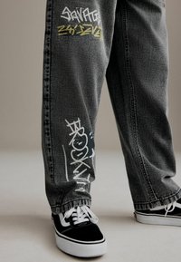 Hallhalli teksad, millel on graafilised prindid, kus on kirjas "SAVAGE" kollase värviga ja "BROOKLYN" valge värviga. Koos mustade Vans'i tossudega.