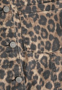 La tela de denim con estampado de leopardo muestra manchas negras sobre un fondo beige. Cuenta con botones de metal y un bolsillo frontal. Superficie texturada evidente.