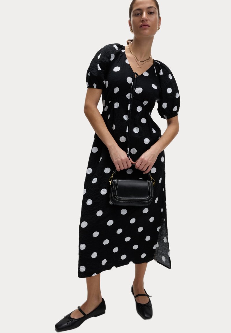 Schwarzes Kleid mit weißen Punkten, kurzen Puffärmeln, V-Ausschnitt und Knopfverschluss. Kombiniert mit einer kleinen schwarzen Handtasche und flachen Schuhen.