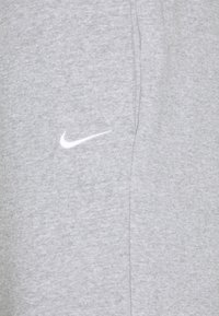 Calças de fato cinzentas feitas de um tecido de algodão misto com uma textura suave. Apresenta um logo da Nike branco perto da cintura e bolsos laterais.