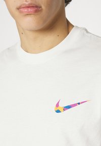 Vit bomull t-shirt med en färgglad Nike-logga i rosa, blått och gult, placerad på vänster bröstområde. Korta ärmar, rund halsringning.