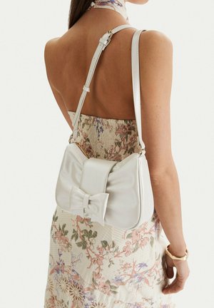 Mujer con un vestido floral sin mangas con escote abierto en la espalda, llevando un bolso de hombro de piel blanca adornado con un gran lazo en la parte trasera.