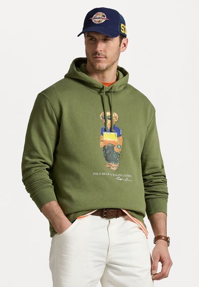 Hanorac de culoare verde măsliniu cu un graphic de urs îmbrăcat într-o cămașă albastră și pantaloni cargo. Textul logo alb în partea de jos. Blugi albi și curea maro.