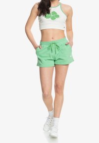 Shorts verts en coton avec une taille élastique et un cordon de serrage. Texture lisse, longueur au genou, avec des poches latérales. Portés avec un haut court blanc.