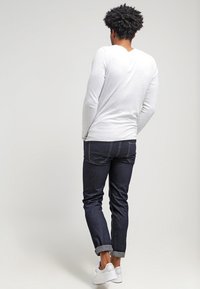 Man med lockigt hår som bär en vit långärmad skjorta, mörka upprullade jeans och vita sneakers, stående vänd bort från kameran mot en enfärgad bakgrund.