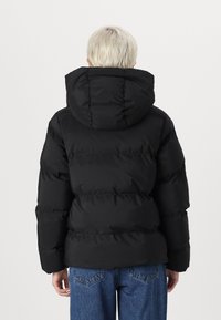 Jachetă puffer neagră cu glugă, având un design matlasat, țesătură texturată și manșete elastice. Purtați deasupra unei perechi de blugi albaștri cu talie înaltă.