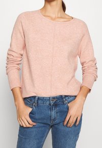 Maglione rosa chiaro, testurizzato, con collo a giro e maniche raglan, abbinato a jeans blu. Dettaglio di cucitura notevole che corre lungo il centro.