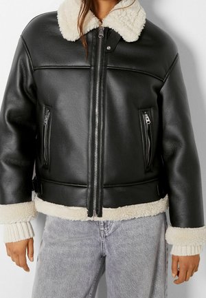 Black leather jacket with cream shearling collar, cuffs, and hem worn over light-wash jeans, featuring front zipper and zippered pockets.