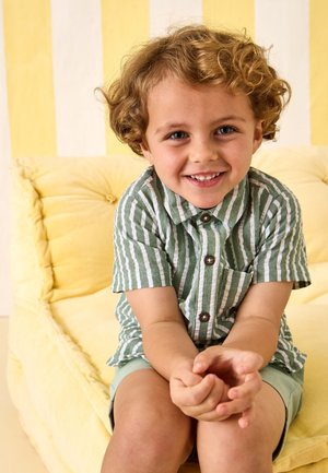 Noor laps lokkis juustega, kandes rohevalge triibulist särki ja pükse, istumas kollasel pehmendusega toolil ja naeratamas.