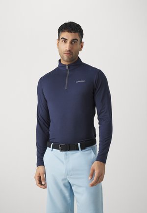 Marineblaue langärmelige Sportoberteil mit einem Viertel-Reißverschluss und dezentem Logo. Getragen mit hellblauen Hosen und einem schwarzen Gürtel in einem schlichten Umfeld.