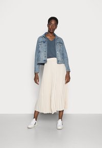 Veste en jean, haut gris en v, jupe midi plissée crème, et baskets blanches avec des accents noirs. La tenue présente une texture en couches.