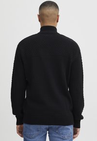 Mann trägt einen schwarzen strukturierten Strickpullover mit hohem Kragen und blaue Jeans, von der einfachen weißen Wand abgewandt.