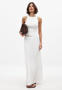 Weißes geripptes Crop-Top, kombiniert mit einem strukturierten weißen Maxi-Rock. Sie hält eine braune Tasche und zeigt ein weiches, leichtes Material sowie ein tailliertes Design.