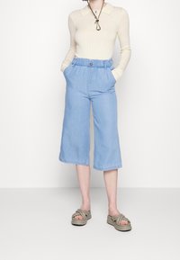 Mujer vistiendo un polo beige de manga larga acanalado, pantalones de pierna ancha cortos de cintura alta en azul claro, y sandalias de plataforma taupe con tiras entrecruzadas.