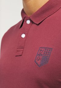 Burgundy polo skjorta med strukturerad stickning, tre knappslås och en marinblå broderad sköldlogga med golfklubbor och stjärnor.