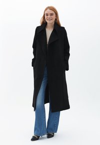 Cappotto nero a maniche lunghe in materiale morbido con colletto ampio, tasche laterali e taglio dritto. Abbinato a jeans a zampa d'elefante blu e tacchi neri.
