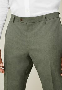 Olive green trousers, made of textured fabric, featuring a button closure and belt loops. Close-fitting cut with pressed creases.