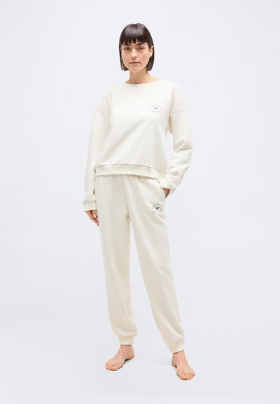 Γυναίκα όρθια, ξυπόλυτη, φοράει εκρού φούτερ και τζόγκερ με τα λογότυπα της Emporio Armani στο στήθος και στον μηρό.