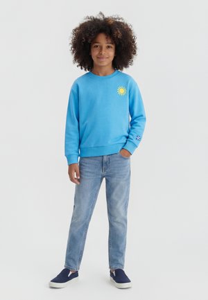 Copil cu păr creț purtând un hanorac albastru cu un motiv galben de soare, blugi deschiși la culoare și pantofi slip-on bleumarin, stând cu o mână în buzunar.