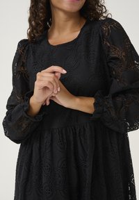 Čierne čipkované šaty s dlhými, nafúknutými rukávmi. Obsahujú kvetinové vzory, okrúhly výstrih a jemné volanty na manžetách.