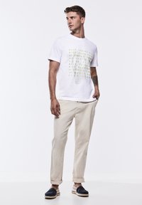 Bílá tričko s krátkým rukávem a zeleným grafickým textem. Kombinováno se světle béžovými kalhotami s ohrnutým rukávem a námořnickými espadrilkami s hnědými podrážkami.