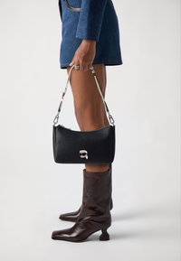 Borsa a tracolla nera con texture, dotata di chiusura a zip, caratterizzata da una tracolla in catena argentata e un piccolo dettaglio con logo. Il modello indossa stivali marroni con tacco e un blazer blu.