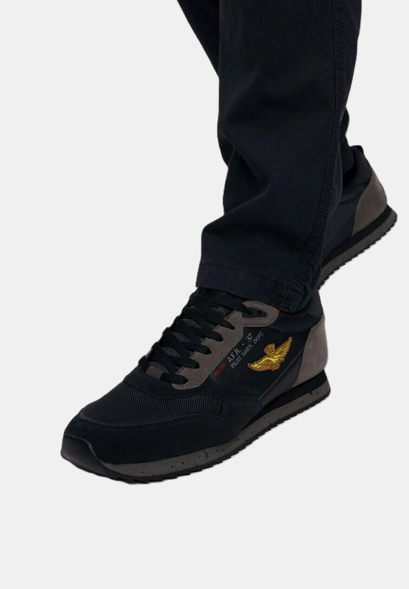 Scarpe da ginnastica nere con accenti grigi, materiali in rete e suede, design con lacci e un logo giallo ricamato sul lato.