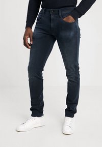 Mies, który ma na sobie ciemnoniebieskie dżinsy w slim fit i białe trampki, z jedną ręką w kieszeni, stojący na jednolitym jasnym tle.