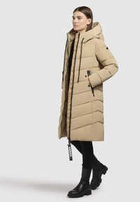 Beige puffatakki hupulla, jossa on viistoviivamalli, etuvetoketju ja mustat yksityiskohdat. Yhdistetty mustiin legginsseihin ja jykeviin mustiin saappaisiin.