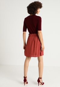 Top in magenta a maglia con maniche corte, abbinato a una gonna color ruggine con vita elasticizzata e scarpe con tacco rosso.