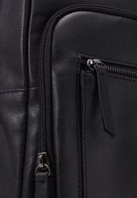 Sac en cuir noir avec une fermeture éclair bien visible, surface texturée et détails de couture discrets, accompagné d'un petit accent en cuir pour le tirette de la fermeture.