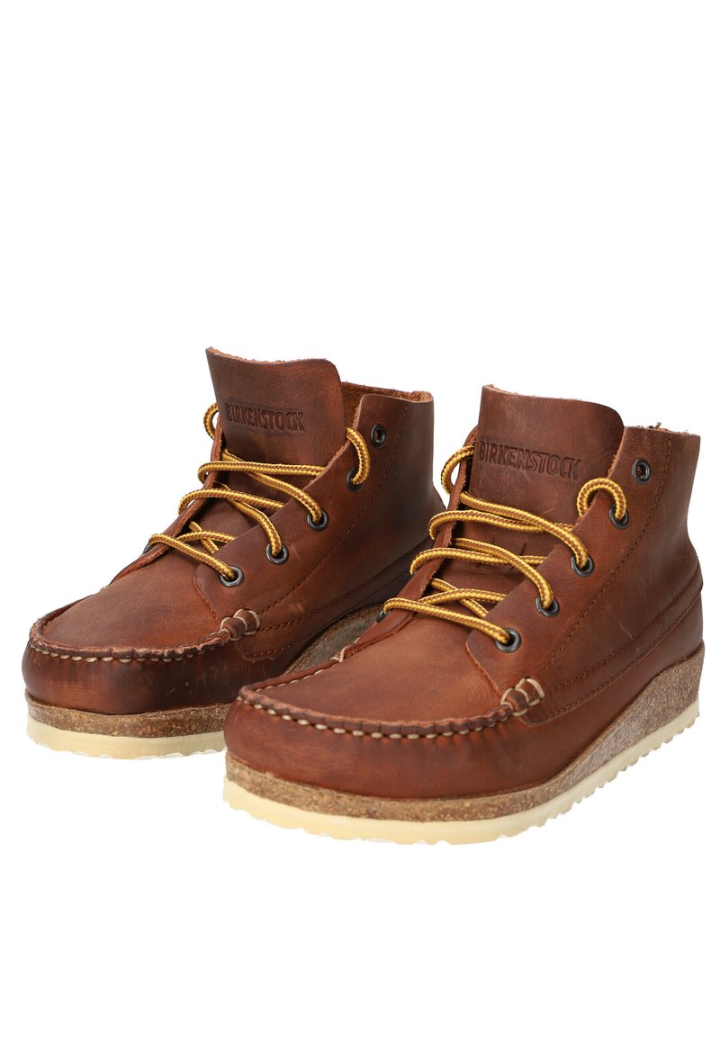 Stivali alla caviglia Birkenstock in pelle marrone con lacci gialli, cuciture moc toe, soletta in sughero e suola in gomma bianca, affiancati su uno sfondo bianco.