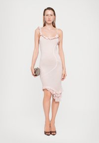 Abito senza maniche di un delicato rosa pallido con spalline arricciate, sagoma aderente, orlo asimmetrico e texture morbida. La modella tiene in mano una piccola borsa metallica.