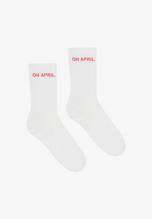 Koronkowy top na ramiączkach w kolorze koralowym z białym logo, zestawiony z szarymi szortami rowerowymi w czarny leopardski wzór. Model nosi białe sneakersy i skarpetki.