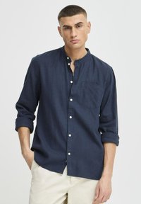 Camisa de lino azul marino con cuello alzado, bolsillo frontal y botones blancos. Presenta mangas enrolladas y un corte relajado.