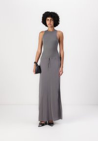Robe maxi gris ajustée avec un col haut et une taille élastique. Le modèle tient un petit sac à main noir, portant des escarpins noirs à bout pointu.